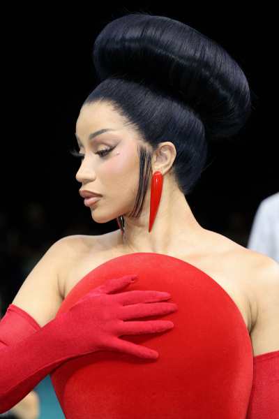 Cardi B appears at a couture fashion show in a red dress gloves and earrings. Her hair is styled in a high circular updo.